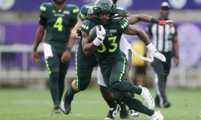 Running back in a green football uniform clutching the ball and sprinting forward while defenders chase on a grassy field.