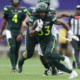 Running back in a green football uniform clutching the ball and sprinting forward while defenders chase on a grassy field.