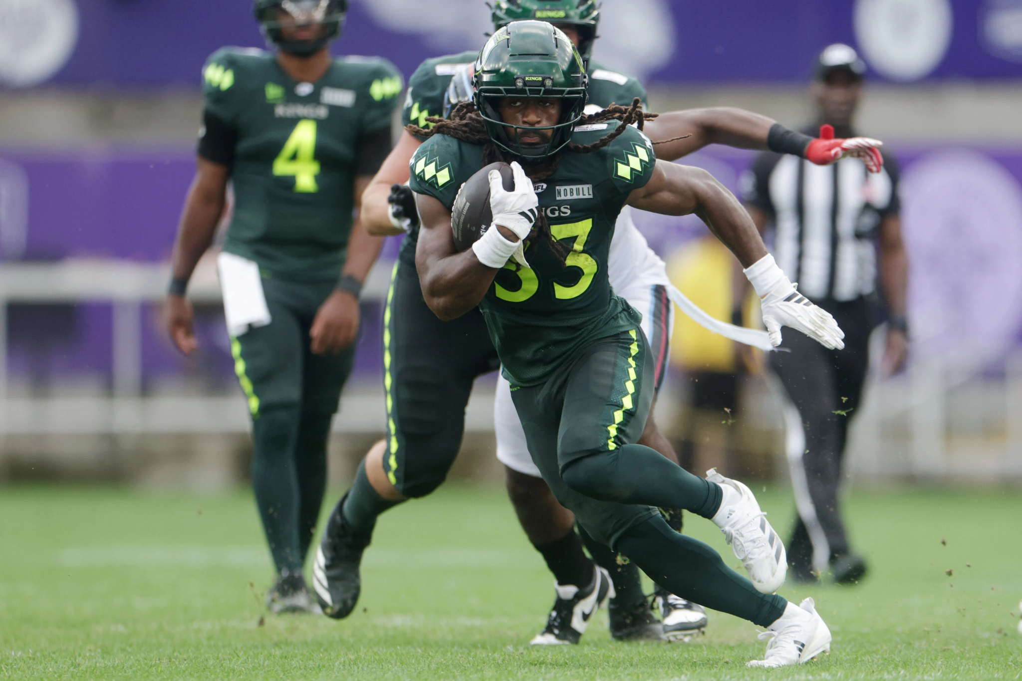 Running back in a green football uniform clutching the ball and sprinting forward while defenders chase on a grassy field.